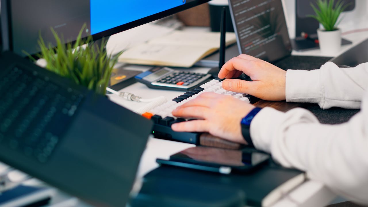 gallery-04 A close-up of a person typing on a keyboard in a modern tech workspace with gadgets and a monitor.